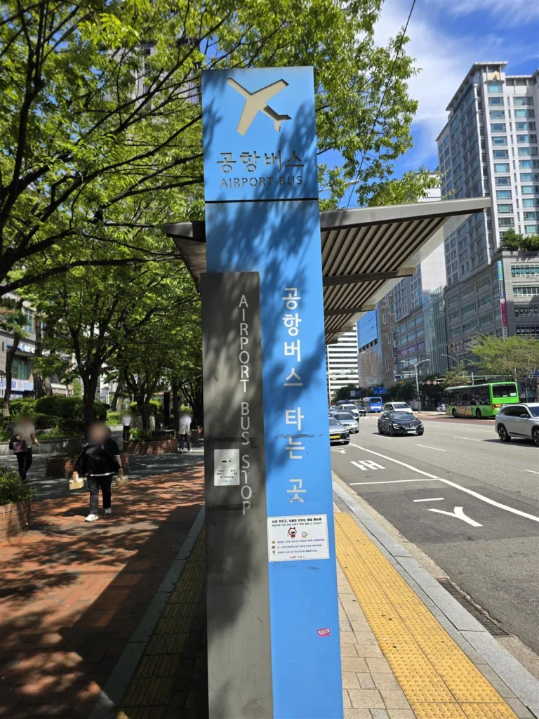 街路沿いにある青いAIRPORT BUS標識と待合屋根。顔はぼかし処理済み